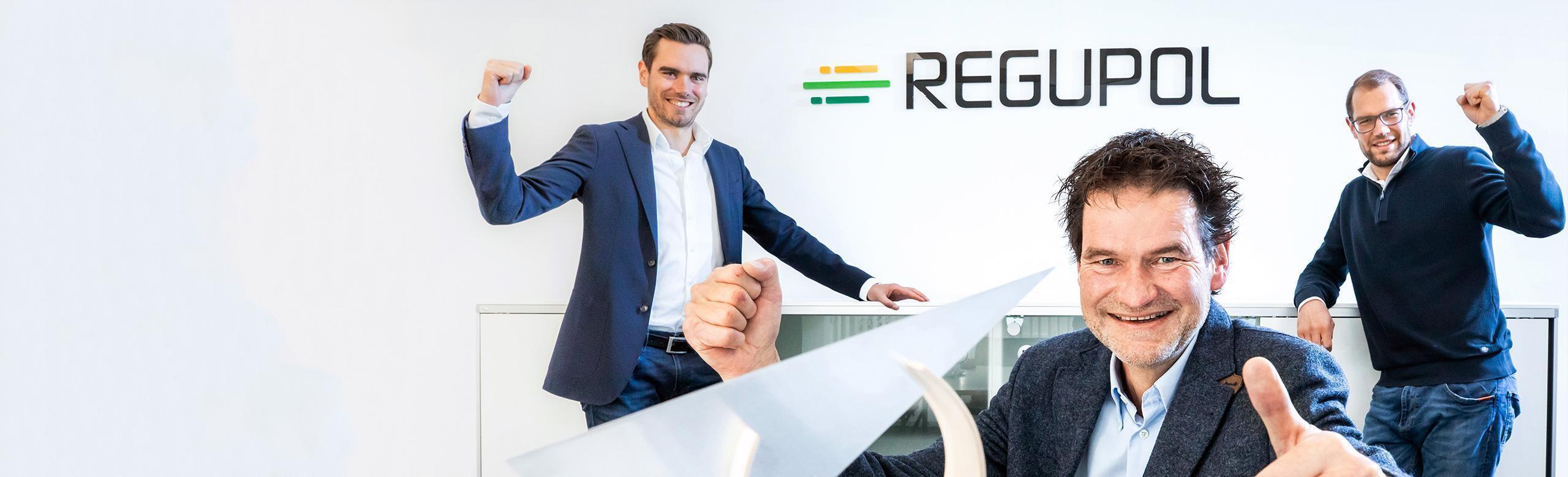 REGUPOL CEOs with Environmental Award – sustainability, recognition, leadership Three men, the CEOs of REGUPOL, stand smiling with raised arms in a bright room in front of the company logo. In the foreground is the Environmental Business Award – a modern, sculptural trophy symbolizing sustainability and achievement.