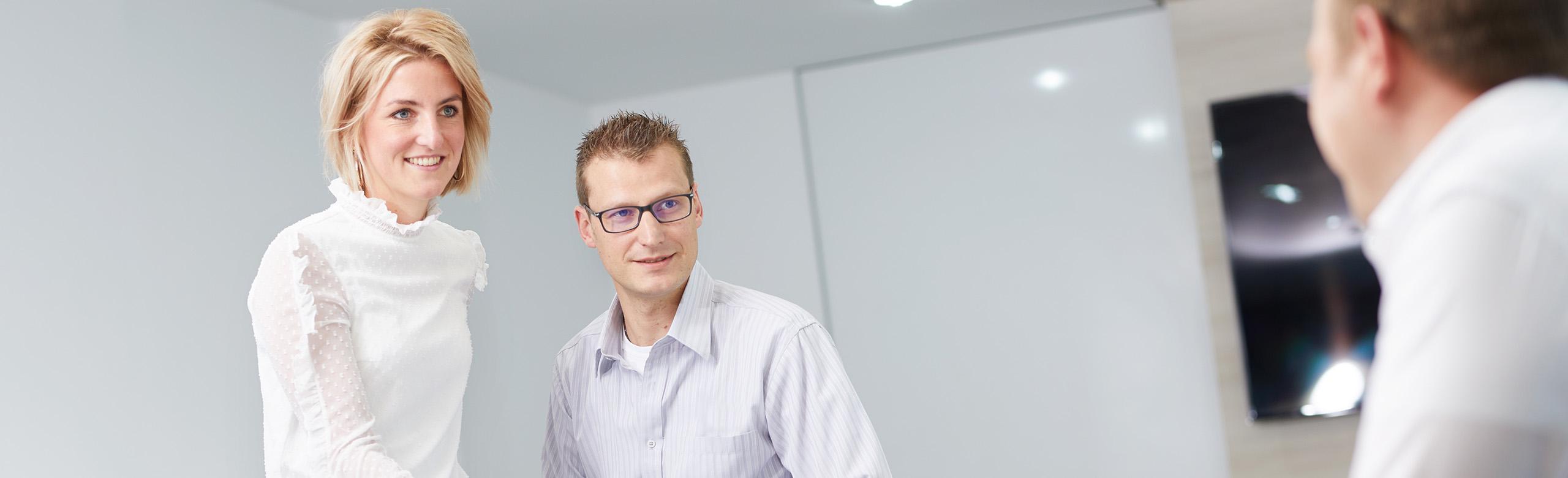Business meeting in office with colleagues in a friendly, professional atmosphere In a modern, bright office, three people are having a conversation. A woman in a white blouse is standing and smiling next to a seated man with glasses. Both are looking toward a third person who is only visible from behind. The atmosphere appears professional and collegial.