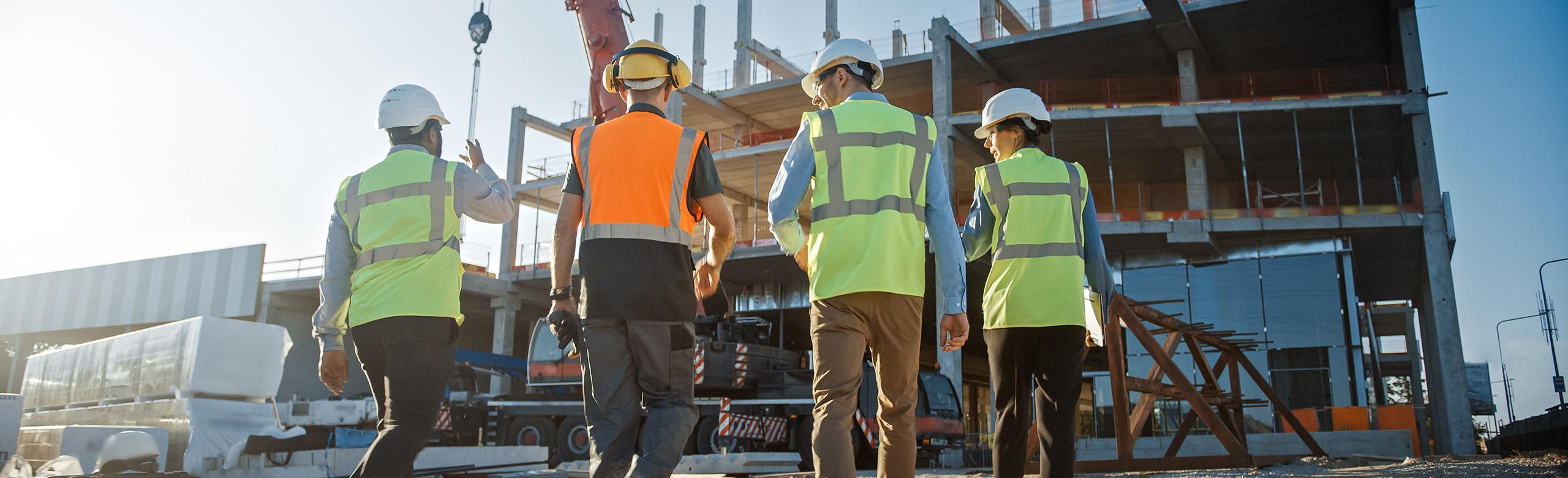 Construction site with team – safety, architecture, planning, sustainable building Four people wearing safety vests and helmets walk across an active construction site. In the background is a partially constructed concrete building, with crane and equipment visible in bright sunlight.