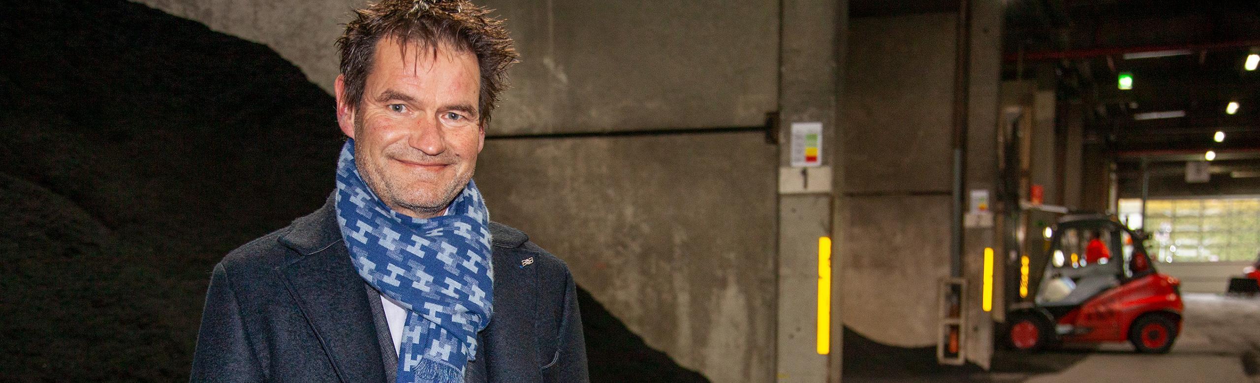 REGUPOL CEO in production – recycling, leadership, industry, sustainability One of REGUPOL’s CEOs stands in an industrial hall wearing dark clothing and a blue patterned scarf. In the background are bulk materials and a red forklift in operation.