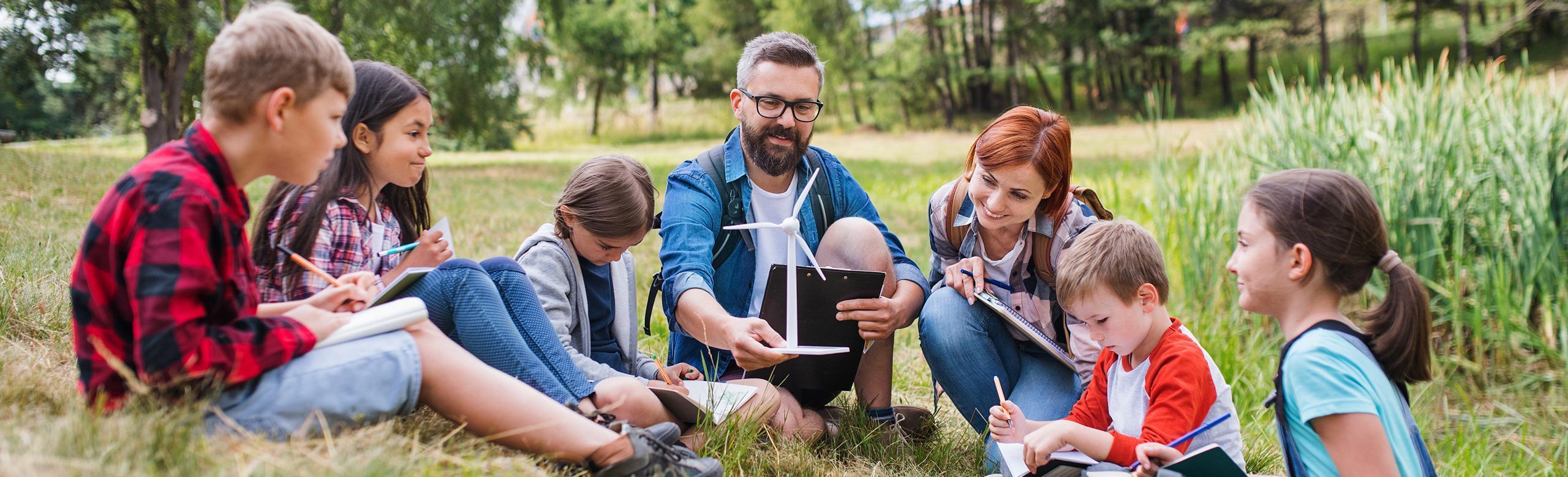 Outdoor environmental learning – kids, wind turbine model, sustainability, nature A group of children sits with two adults in a circle on a meadow. They are learning outdoors, using notebooks and a wind turbine model. Trees and reeds can be seen in the background.